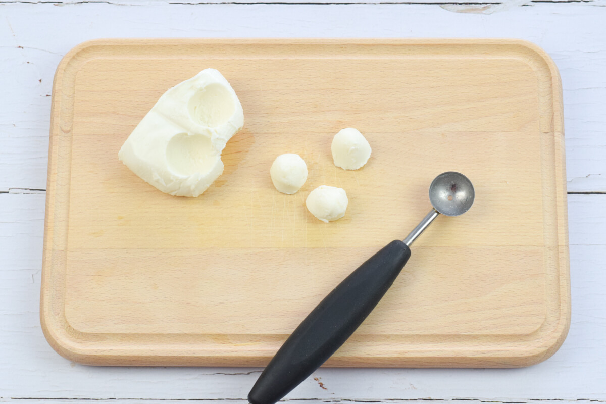 A wooden chopping board with a melon baller and 3 small balls of mozzarella next to a halved ball of mozzarella 